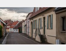 Schmale, gepflasterte Straße in einem Wohngebiet mit Reihenhäusern. Die Gebäude haben helle Fassaden, Fensterläden in Pastellfarben und rote Ziegeldächer; der Himmel ist bewölkt.