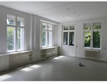 Heller, leerer Raum mit großen weißen Fenstern und Glastür, die nach draußen zu einer grünen Gartenszenerie führen. Entlang der Wände befinden sich weiße Heizkörper und integrierte Regale.