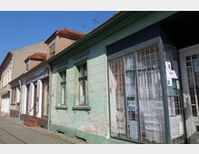 Eine Reihe von älteren Häusern mit bröckelnder Fassadenfarbe in einer gepflasterten Straße. Ein kleines Schaufenster mit Gitter, dahinter Gardinen, befindet sich rechts im Bild.