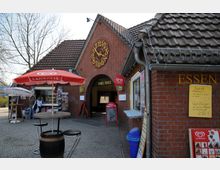 Backsteingebäude mit einem großen Ziffernblatt auf dem Giebel und einem Durchgang mit der Aufschrift "Thiel Platz". Vor dem Gebäude befinden sich ein Verkaufsstand mit roten Sonnenschirmen, Tischen und Stühlen sowie eine Speisekarte an der rechten Wand.