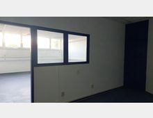 Leerer Raum mit weißen Wänden, blauer Fensterrahmung und eingebauten Glasfenstern, die in einen angrenzenden, hell erleuchteten Raum blicken. Darüber hinaus sind Steckdosen entlang der Wand und eine abgehängte Rasterdecke sichtbar.