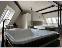 Modernes Dachgeschoss-Schlafzimmer mit freistehender Whirlpool-Badewanne im Vordergrund, schrägen Dachfenstern und sichtbaren schwarzen Deckenbalken. Im Hintergrund stehen ein großes Bett mit Nachttischen sowie ein Schrank; durch die Fenster ist ein Blick über die Stadt möglich.