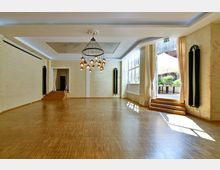 Heller, leerer Saal mit hohen Decken, großen Fenstern auf der rechten Seite, einem glänzenden Holzfußboden und einer zentralen Hängelampe. Eine geöffnete Tür mit Treppenstufen führt nach draußen zu einer Terrasse mit Sonnenschirm und Pflanzen.