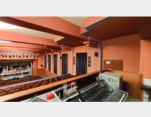 Blick in einen roten Theatersaal mit Reihen aus braunen Sitzen. Im Vordergrund befindet sich ein Technikpult, im Hintergrund sieht man eine Bühne mit Beleuchtung und Bühnenelementen.