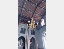 Innenraum einer Kirche mit einem dunklen, reich verzierten Holzdach und gotischen Spitzbogenfenstern. Ein großer, goldener Kronleuchter hängt zentral, und im Hintergrund ist eine Orgel mit goldenen Details sowie eine Treppe zu sehen.