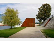 Grünfläche an einem Ufer mit einem kunstvollen, rostfarbenen Metallobjekt. Links steht ein Baum, im Hintergrund sind ein Wasserweg, moderne Gebäude und ein Boot zu sehen; rechts verläuft eine steinerne Uferbefestigung mit einer Schräge.
