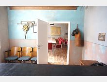 Ein Blick in einen Raum mit hellblau-rosa gestrichenen Wänden, Holzboden und hölzernen Stühlen mit gepolsterten Sitzflächen. Durch eine offene Tür führt der Blick in einen angrenzenden Konferenzraum mit einem großen Tisch, rotem Stuhlbezug und einer Karte an der grauen Betonwand. Boxhandschuhe und ein Boxsack hängen an der Wand neben der Tür.