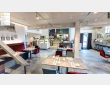 Helles Restaurant mit modernem Interieur, ausgestattet mit rot- und blau gepolsterten Stühlen und gedeckten Tischen. Im Hintergrund befinden sich eine offene Theke mit Pflanzen und Regalen sowie ein Ausgang mit einer halbkreisbogenförmigen Glastür.