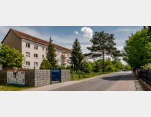 Wohnstraße mit einem mehrstöckigen Wohngebäude links, dessen Fassade beige ist und ein rotes Schrägdach hat. Entlang der Straße befinden sich Gehwege, Bäume und Grünflächen, die eine ruhige, begrünte Umgebung schaffen.