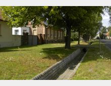 Straßenszene in einer ländlichen Gegend mit Baumalleen, einem Ziegelgebäude im Hintergrund und einem kleinen, gepflasterten Wassergraben entlang eines Gehwegs. Rechts befindet sich ein Bushaltestellenschild neben einer asphaltierten Straße.