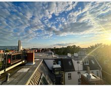 Dächer einer Stadt bei Sonnenuntergang mit teils bewölktem Himmel und Blick auf moderne und historische Gebäude sowie vereinzelte Bäume im Hintergrund. Eine Dachterrasse mit Metallgeländer und gemauerten Elementen ist im Vordergrund zu sehen.