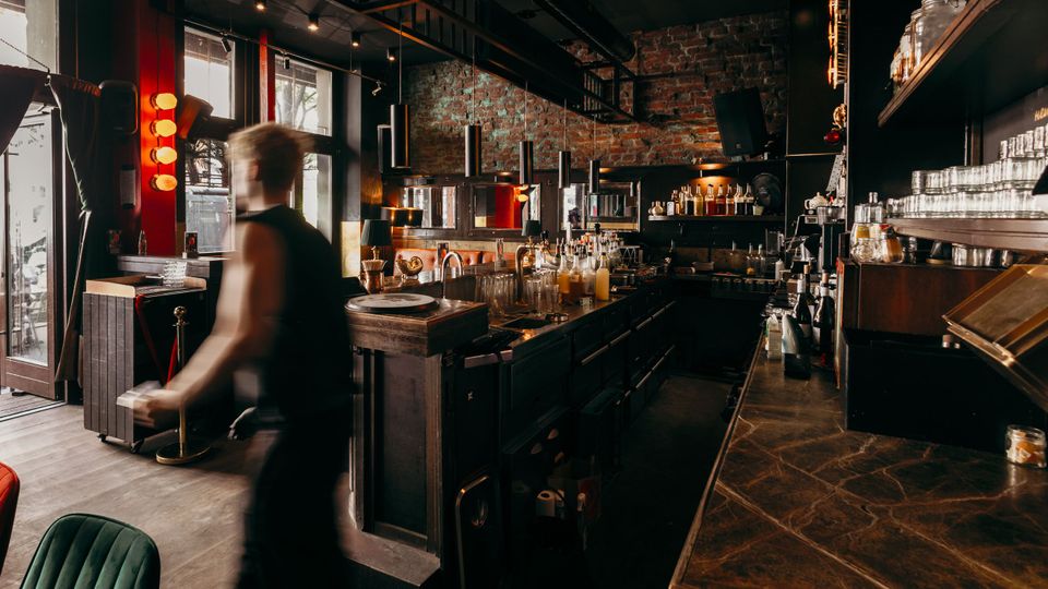 Innenraum einer Bar mit langer dunkler Theke, Regalen voller Gläser und Flaschen sowie warmem Licht. Backsteinwand, schwarze Decke mit Industrieelementen und große Fenster zur Straße prägen den Raum.