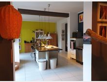 Moderne Wohnküche mit zentraler Kücheninsel, Edelstahlspüle und drei Hängelampen. Farbige Wände in Gelb und Dunkelblau, dekorative Kunstwerke und Holzmöbel prägen den Raum; links ist ein oranger Lampion zu sehen.