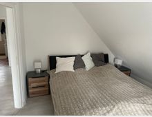 Schlafzimmer mit einem Doppelbett unter einer Dachschräge, flankiert von zwei Nachttischen mit Lampen. Im Hintergrund führt eine offene Tür zu einem angrenzenden Raum mit Fliesenboden.