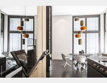 Moderne Innenraumaufnahme einer offenen Küche mit hellen Holz- und schwarzen Elementen, daneben ein Essbereich mit rundem Tisch und weißen Stühlen vor großen Fenstern mit Jalousien. Eine minimalistische Pendelleuchte mit farblich gemischten, organisch geformten Lampenschirmen hängt von der Decke.