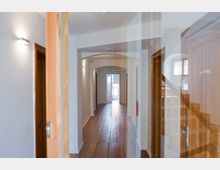 Ein Innenraum mit einem schmalen Flur, Holzboden und weiß gestrichenen Wänden. Der Flur führt zu einer geöffneten Glastür am Ende, auf beiden Seiten befinden sich Türen mit Holzrahmen, und rechts ist ein Teil einer Holztreppe sichtbar.