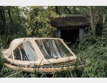 Ein verlassenes Boot mit beigem Verdeck liegt von dichtem Grün und hohem Gras umgeben vor einer kleinen, verwitterten Holzhütte im Wald. Die Umgebung ist naturbelassen mit vielen Bäumen und Buschwerk.