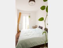 Helles Schlafzimmer mit einem großen Bett, einer grünlichen Tagesdecke und Holzboden. Im Raum befinden sich ein geflochtener Stuhl, eine gelbe Vorhangschiene neben einer Balkontür sowie Pflanzen als Dekoration.