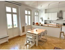 Heller Wohnküchenbereich mit hohen, bogenförmigen Fenstern, Fischgrät-Parkettboden und einem Holztisch mit mehreren Stühlen im Vordergrund. Im Hintergrund eine moderne weiße Küchenzeile mit Fliesenrückwand, Dunstabzugshaube und pendelnden Deckenlampen über einer langen Arbeitsfläche.