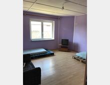 Ein spärlich eingerichtetes Zimmer mit lila Wänden und Holzfußboden. Es enthält ein Fenster mit Blick auf eine Mauer, eine Matratze auf Paletten, einen kleinen Röhrenfernseher auf einem Holzgestell und ein graues Sofa.