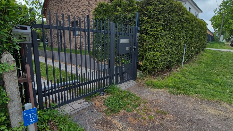 Einfamilienhaus aus Ziegelmauerwerk mit steilem, dunkel gedecktem Satteldach und kleiner Dachantenne, hinter einem hohen schwarzen Metallzaun mit Tor. Rechts verläuft eine Hecke entlang des Grundstücks, daneben ein schmaler Weg und Grünstreifen unter bewölktem Himmel.