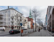 Straßenszene in einer Stadt mit Wohn- und Geschäftsgebäuden, darunter ein modernes weißes Gebäude mit Balkonen und eine rote Backsteinkirche mit grünem Spitzdach im Hintergrund. Auf der Straße fahren Autos, ein Fahrradfahrer ist sichtbar, und am Gehweg stehen Fußgänger sowie eine Bushaltestelle.