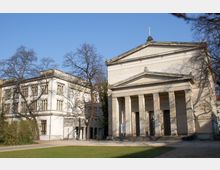 Historisches Gebäudeensemble mit klassizistischer Kirche rechts, erkennbar an ihrem Säulenvorbau und Giebeldreieck, sowie einem angrenzenden weißen, rechteckigen Gebäude mit großen Fenstern. Im Vordergrund befindet sich eine Rasenfläche, umgeben von laublosen Bäumen unter klarem, blauem Himmel.