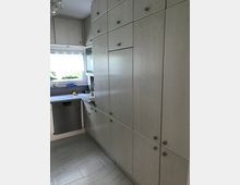 Helle Küche mit grauen Holzschränken, einer Arbeitsplatte aus Stein und einem eingebauten Geschirrspüler. Rechts stehen hohe Schränke, links führt ein Fenster mit Blick ins Freie Tageslicht herein.