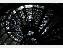 Metallene Wendeltreppe mit durchbrochenem Gittermuster, von unten fotografiert. Im Hintergrund sind Glas- und Metallstrukturen eines modernen Gebäudes sichtbar.