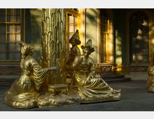 Detailansicht des Chinesischen Hauses im Park Sanssouci, Potsdam, mit vergoldeten Skulpturen in orientalischer Kleidung um eine verzierte Säule. Im Hintergrund sind grüne Wände mit goldgerahmten Fenstern sichtbar.