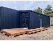 Modernes, dunkel verkleidetes Holzhaus mit flach geneigtem Dach und großen Glasfronten, davor eine breite Holzterrasse mit Stufen. Das Gebäude steht auf sandigem Boden am Rand eines bewaldeten Areals.