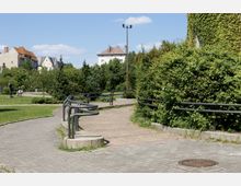 Ein Park mit gepflastertem Gehweg und geschwungener Metallgeländerführung, umgeben von dichter grüner Vegetation. Im Hintergrund sind Wohnhäuser und eine Statue sichtbar.