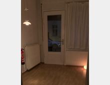 Ein kleiner Raum mit hellem Laminatboden, einer Fenstertür mit Glas, die nach draußen führt, und einem weißen Heizkörper auf beiden Seiten. Links hängt eine kleine Deckenlampe; rechts steht eine leuchtende Kugel auf dem Boden. Ein Feuerlöscher ist an der linken Wand befestigt.