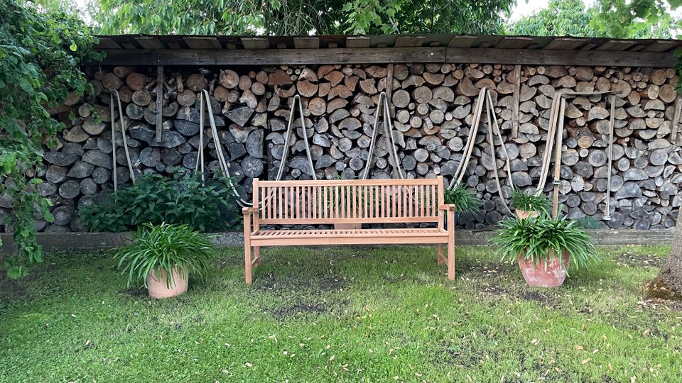 Holzbank auf einer Rasenfläche vor einem überdachten Holzlager, in dem gestapelte Baumstämme und mehrere an der Wand hängende Äxte zu sehen sind. Daneben stehen zwei Pflanzkübel mit grünen Pflanzen, umgeben von Bäumen und Sträuchern.