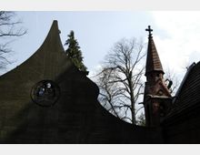 Eine Kirche mit einer geschwungenen, dunklen Fassade, die ein rundes Kirchenfenster mit Glasdetails enthält, und einem Backsteinturm mit spitzem Dach und Kreuz. Im Hintergrund ragen kahle Bäume und ein Nadelbaum in den Himmel.