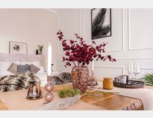 Ein helles Schlafzimmer mit einem sorgfältig dekorierten Holztisch im Vordergrund, auf dem eine rosa Glasvase mit roten Zweigen, zwei Kerzenhalter, eine kleine Pflanze und ein Tablett mit Gläsern und Tassen stehen. Im Hintergrund befindet sich ein Bett mit Kissen und einer Felldecke, weiße Wände mit Wandvertäfelung und minimalistischen Kunstdrucken.