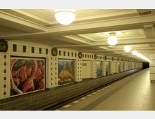 U-Bahn-Station mit hell beleuchtetem Bahnsteig, verziert mit bunten Wandmalereien in rechteckigen Rahmen entlang der Wand; die Decke ist mit dekorativen Reliefmustern und kugelförmigen Lampen ausgestattet.