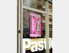 Ein Schaufenster mit einer großen weißen Aufschrift „Pastiche“ und einem rosa Poster, das Informationen über ein Festival am 29. und 30. März anzeigt. Im Hintergrund spiegeln sich Bäume und Gebäude.