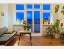 Wohnzimmer mit Holzboden, einer grauen Couch mit gelbem Kissen links, einem kleinen Holztisch in der Mitte und mehreren Topfpflanzen auf einem Fensterbrett und daneben. Große Fenster mit Blick auf einen blauen Abendhimmel und eine Terrasse im Hintergrund.