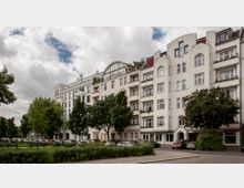 Straßenansicht mit mehrstöckigen Wohnhäusern im Altbaustil, weiß gestrichener Fassade und mehreren Balkonen. Vor den Gebäuden befinden sich geparkte Autos, Bäume und eine kleine Grünanlage entlang des Gehwegs.