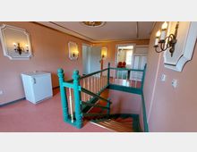 Innenraum eines Treppenhauses mit rosa gestrichenen Wänden, dekorativen Wandlampen und grünem Holzgeländer. Im Hintergrund befindet sich eine offene Tür, die in einen Raum mit roten Stühlen und einem Kronleuchter führt.