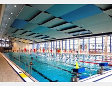 Hallenbad mit einem großen Schwimmbecken, das durch rote und blaue Bahnenleinen unterteilt ist. Große Fensterfronten lassen Tageslicht herein, während die Decke mit blauen und grauen Schalldämmplatten gestaltet ist; am Beckenrand befinden sich Startblöcke und einige Personen.