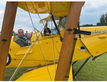 Ein gelbes Doppeldeckerflugzeug mit Holzstreben und offenem Cockpit, in dem ein Pilot in grüner Uniform sitzt. Im Hintergrund sind einige Zuschauer auf einer Wiese sowie Bäume und ein bewölkter Himmel sichtbar.
