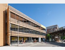 Mehrstöckiges modernes Parkhaus mit Fassade aus horizontalen Holzlamellen und großen Fensterflächen. Im Vordergrund fahren zwei Personen auf roten Fahrrädern vorbei, rechts führt eine erhöhte Bahngleiskonstruktion entlang.