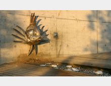 Metallskulptur eines Fisches mit stacheligen Flossen an einer Betonwand, daneben ein Holzsteg und darunter ein flacher Wasserlauf mit weißen Steinen.