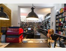 Ein gemütlicher Buchladen mit integriertem Café, erkennbar an der roten Kaffeemaschine auf dem Tresen im Vordergrund. Im Hintergrund sind hohe Regale voller Bücher und andere Verkaufsartikel sowie eine Theke mit Kasse zu sehen, beleuchtet von großen, hängenden Lampen.