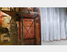 Bühnenbereich hinter den Kulissen mit einer Holzkonstruktion, Beschilderung "Platz für die Feuerwehr" und schwarzen Metallstreben. Rechts im Bild ist ein geschlossener, grauer Bühnenvorhang sichtbar.