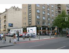 Straßenszene mit einem mehrstöckigen Wohnhaus und Geschäften im Erdgeschoss, darunter ein Café mit Aufschrift „Balzac Coffee“. Vor dem Gebäude geparkte Autos, Fahrräder, Gehweg mit Fußgängern und Straßenschilder, sowie Sitzplätze des Cafés mit Sonnenschirmen.