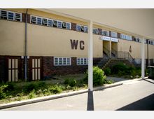 Außenansicht eines öffentlichen Toilettengebäudes mit gelber Fassade und roten Klinkerelementen. Schilder für "WC Damen" und "WC Herren" sind über einer Treppe sichtbar, daneben sind getrennte Eingänge und große Fensterreihen zu erkennen.