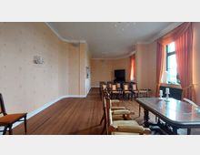 Ein eleganter Raum mit hellorangefarbenen tapezierten Wänden, dekorativen Kronleuchtern und einem Holzboden. Der Raum ist mit einer langen Tafel und Stühlen ausgestattet, im Hintergrund stehen weitere Stuhlreihen sowie ein dunkler Holzschrank. Große Fenster mit orangefarbenen Vorhängen lassen Tageslicht herein.